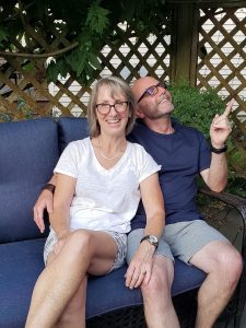 Sue and Stu sitting outside. Stu is happily pointing up to the sun on the roof. More sun means more electricity is produced.